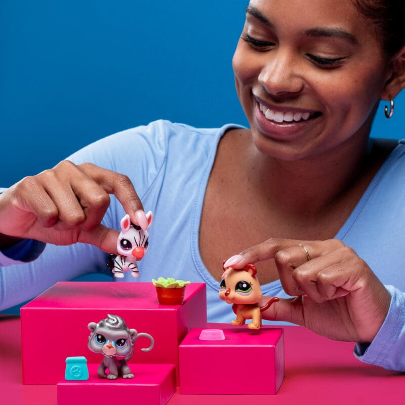 621_Image3 A person with a joyful expression is playing with three toy animals on pink blocks against a blue background. The toys include a zebra, a lion, and a monkey. The person, wearing a light blue top, holds the zebra and lion toys.